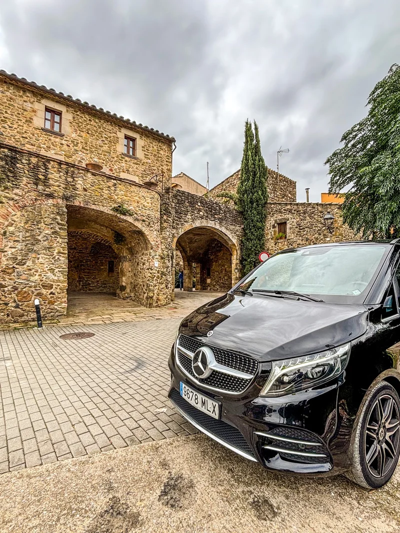 Vehículo negro de chófer privado estacionado frente a los arcos de piedra del casco medieval de Monells, en el Empordà